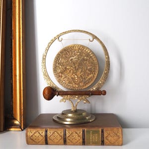 May include: A brass gong suspended within a circular frame, featuring intricate floral engravings on the disc and base. A wooden mallet rests horizontally across the front of the stand. The gong is positioned on top of a rectangular, gold-spined book.