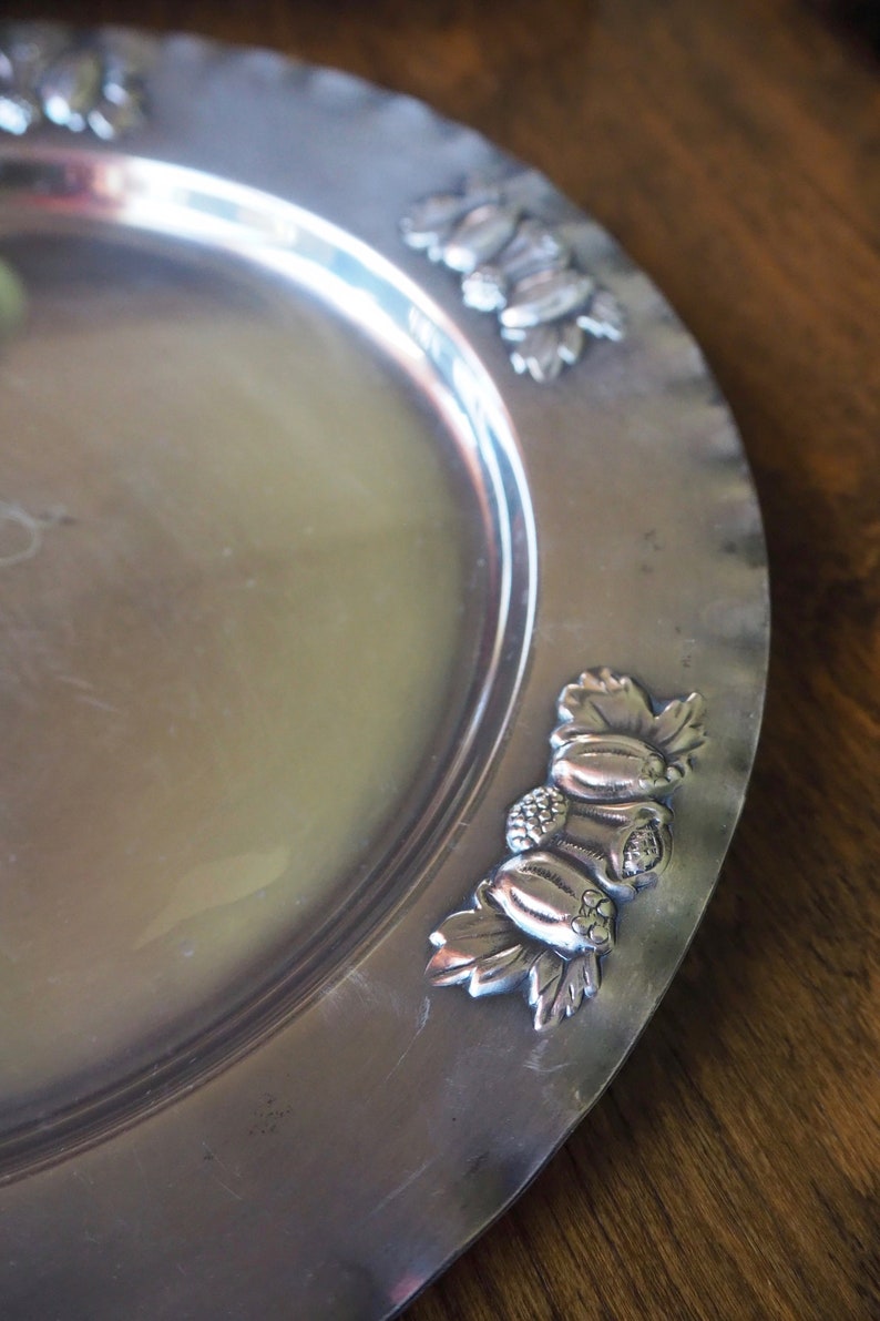 May include: A silver serving platter with a decorative embossed design of leaves and fruit around the edge.