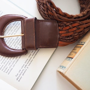 May include: A brown leather belt with a woven design and a large, brown leather buckle. The belt is lying on a book with white pages.