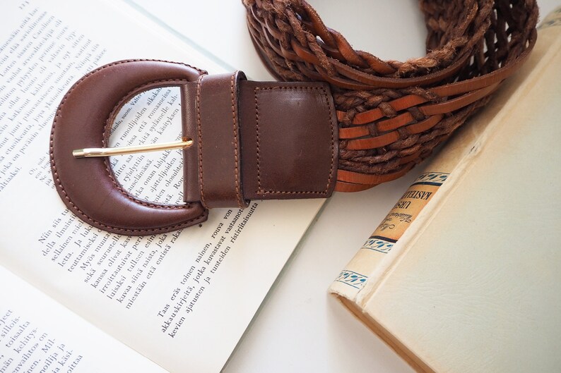 May include: A brown leather belt with a woven design and a large, brown leather buckle. The belt is lying on a book with white pages.