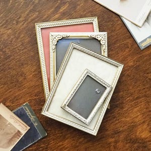 Puede incluir: Un conjunto de cuatro marcos de fotos rectangulares con bordes metálicos dorados, esquinas de filigrana decorativa y bordes texturizados. Los marcos están superpuestos y muestran paspartús en colores coral, gris y crema.