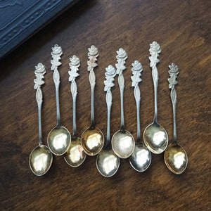 Peut inclure: Ensemble de dix cuillères à café en argent avec des manches en forme de rose. Les cuillères ont une finition dorée et sont disposées en rangée sur une surface en bois.