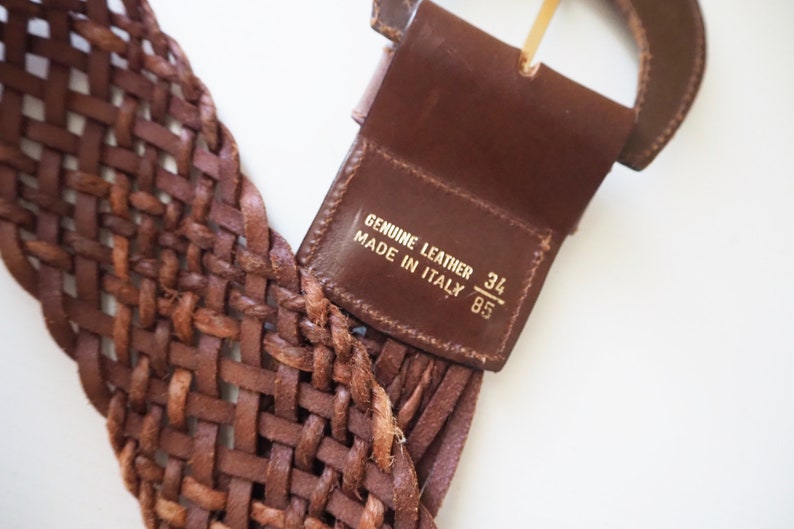 May include: A brown leather belt with a woven design and a gold buckle. The belt has a label that reads "Genuine Leather Made in Italy 34/85".