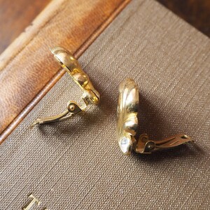 May include: A pair of gold-tone clip-on earrings with a floral design. The earrings are closed and lying on a brown surface.