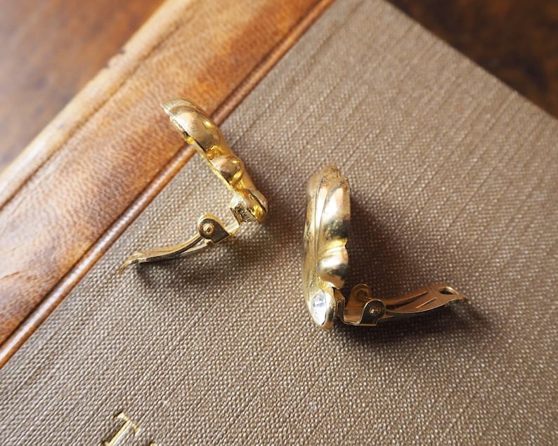 May include: A pair of gold-tone clip-on earrings with a floral design. The earrings are closed and lying on a brown surface.