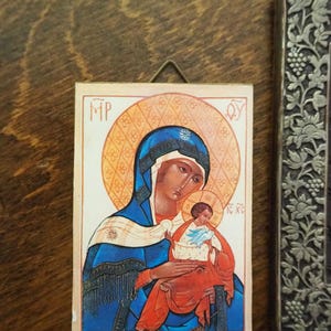 Pu&ograve; includere: Un'icona religiosa che raffigura la Vergine Maria che tiene in braccio il bambino Ges&ugrave;. L'icona &egrave; dipinta in uno stile tradizionale con accenti dorati e uno sfondo bianco. Il testo "MP OY IC XC" &egrave; visibile sull'icona.
