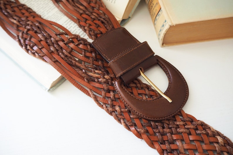 May include: A brown leather belt with a woven design and a large, brown leather buckle with a gold metal clasp.