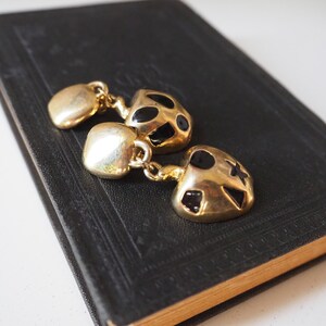 May include: A pair of gold-tone earrings with black enamel accents. The earrings are shaped like a heart and have a dangling charm.
