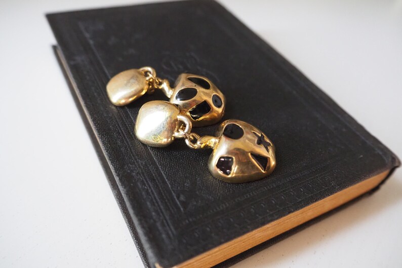 May include: A pair of gold-tone earrings with black enamel accents. The earrings are shaped like a heart and have a dangling charm.