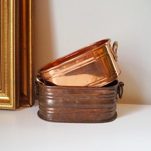 May include: Two copper oval planters, one nested inside the other. The top planter has a handle on one side. Both planters have a shiny, metallic finish.