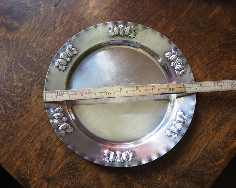 May include: A silver serving tray with a decorative border of embossed leaves and berries. The tray is 8 inches in diameter.