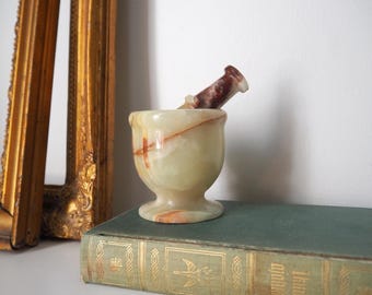May include: A small, pale green onyx mortar and pestle set. The mortar has a rounded bowl shape with a pedestal base. The pestle is a darker brown color. The set is displayed on a vintage book with gold lettering.
