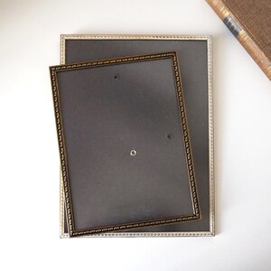 Puede incluir: Dos marcos de fotos rectangulares. El marco más grande tiene un borde plateado con un diseño de cuentas, y el marco más pequeño tiene un borde dorado con un diseño de cuentas similar. Ambos marcos tienen un respaldo gris oscuro.