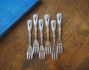May include: Set of six silver-toned forks with ornate rose and leaf designs. The forks are arranged in a fan shape on a wooden surface.