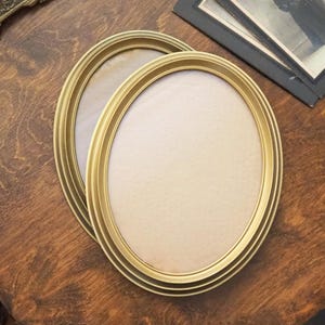 Peut inclure: Deux cadres photo ovales dorés avec un design à plusieurs couches. Les cadres sont vides, prêts à accueillir des photos. Ils reposent sur une surface en bois sombre, avec une esthétique vintage. Un petit objet en métal orné et une pile de vieilles photos sont en arrière-plan.