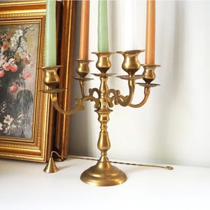 May include: A gold-toned metal candelabra with five candle holders. The candelabra has a decorative design and is holding four candles, two are peach and two are green.