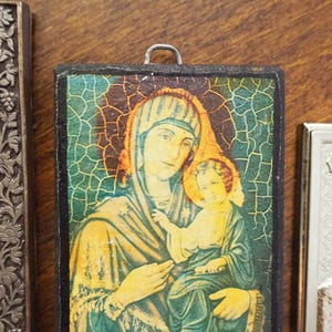 Pu&ograve; includere: Una piccola icona incorniciata in legno che raffigura una donna in un abito blu e verde che tiene un bambino. L'icona ha un effetto di vetro screpolato e uno sfondo dorato.