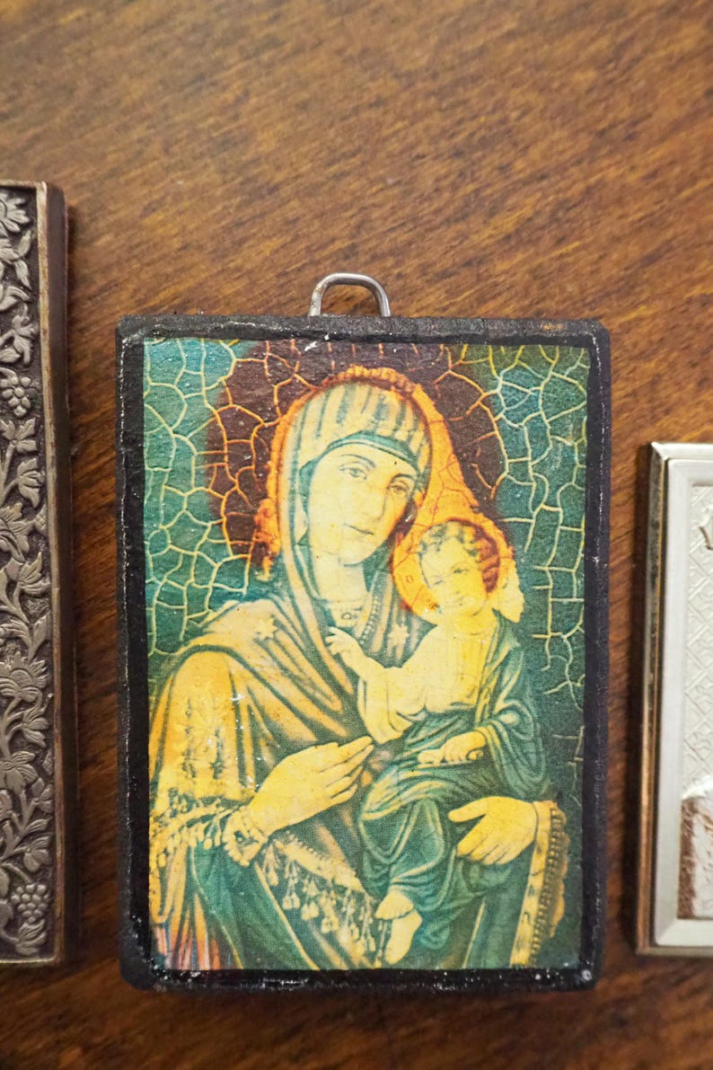 Pu&ograve; includere: Una piccola icona incorniciata in legno che raffigura una donna in un abito blu e verde che tiene un bambino. L'icona ha un effetto di vetro screpolato e uno sfondo dorato.