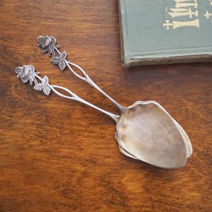 Könnte beinhalten: Silberfarbene Servierzange mit Rosen- und Blattdetails an den Griffen. Die Zange hat einen gewellten Rand am Servierende. Der Hintergrund ist eine dunkle Holzoberfläche, mit einem Vintage-Buch in der oberen rechten Ecke.