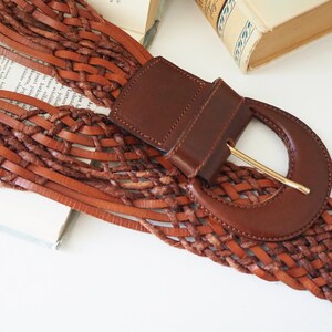 May include: A brown leather belt with a woven design and a large, brown leather buckle with a gold metal clasp.