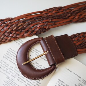 May include: A brown leather belt with a woven design and a large, brown, oval-shaped buckle.
