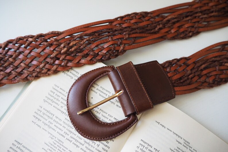 May include: A brown leather belt with a woven design and a large, brown, oval-shaped buckle.