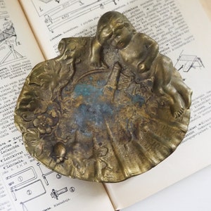 May include: A vintage brass ashtray with an ornate design featuring a cherub figure and floral motifs. The ashtray has a worn, aged patina and is a unique decorative piece.