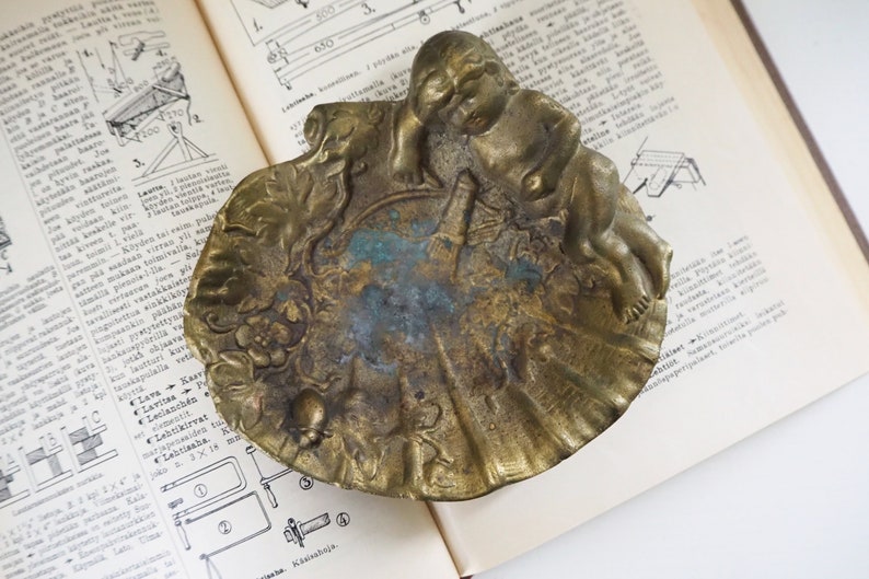 May include: A vintage brass ashtray with an ornate design featuring a cherub figure and floral motifs. The ashtray has a worn, aged patina and is a unique decorative piece.