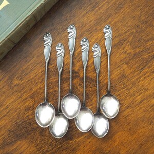 May include: A set of six silver-toned metal spoons with long, slender handles. Each handle features a distinct, raised spiral or floral-like motif at the top. The spoons have polished, oval-shaped bowls and a smooth, metallic finish throughout.