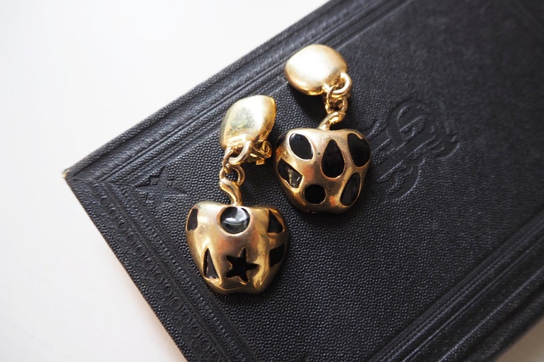 May include: A pair of gold tone clip-on earrings with black and gold heart-shaped charms. The charms have a polka dot pattern and a star design.