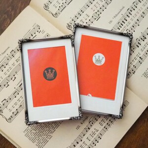 Puede incluir: Dos cuadros enmarcados con marcos plateados, cada uno con un fondo naranja brillante y un emblema de corona. Los marcos descansan sobre un fondo de partituras antiguas, creando una exhibición clásica y artística.