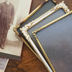 May include: Three antique picture frames with ornate gold and white detailing. The frames are empty, with a dark gray backing. A vintage photograph of a man in a suit is partially visible in the background.