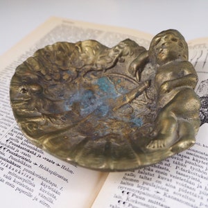 May include: A vintage brass trinket dish with an ornate design featuring a cherub figure. The dish has a worn, aged patina and is perfect for holding small items.
