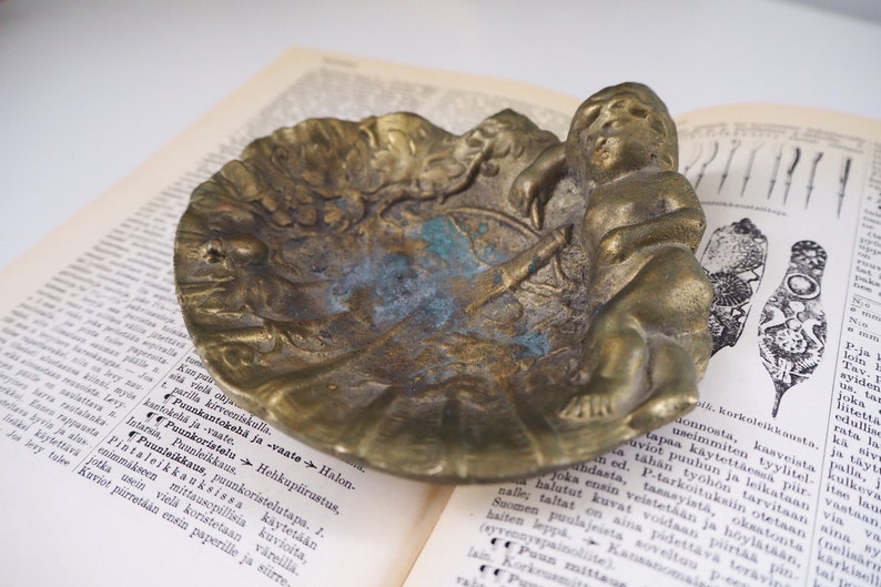 May include: A vintage brass trinket dish with an ornate design featuring a cherub figure. The dish has a worn, aged patina and is perfect for holding small items.