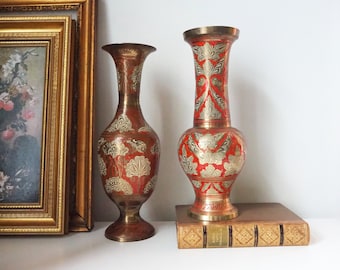 May include: Two decorative brass vases with intricate red and gold designs. The vases are on a shelf with a book.