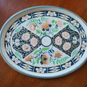Puede incluir: Una bandeja de servir ovalada de metal con fondo blanco y un dise&ntilde;o floral en tonos azules, verdes, rosas y naranjas. El dise&ntilde;o presenta flores, hojas y espirales estilizadas.
