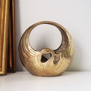 Puede incluir: Un jarrón de metal dorado con forma de cisne con las alas extendidas. El jarrón tiene una abertura circular en la parte superior y está diseñado para contener flores u otros artículos decorativos.