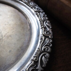 May include: A silver serving tray with an ornate, embossed design around the edge. The tray is circular and has a smooth, reflective surface.