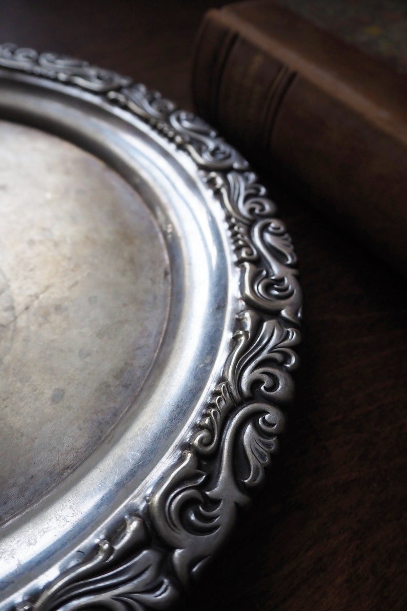 May include: A silver serving tray with an ornate, embossed design around the edge. The tray is circular and has a smooth, reflective surface.