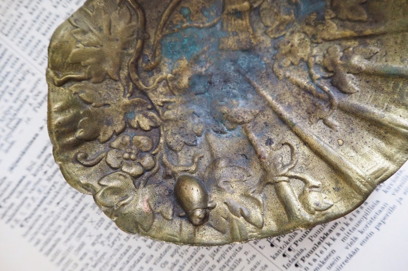 May include: A vintage brass trinket dish with an ornate design featuring leaves, flowers, and a small snail. The dish has a worn, aged patina.
