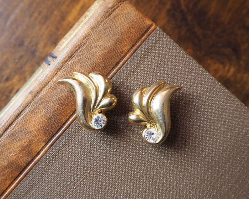 May include: A pair of gold-tone earrings with a leaf design and a clear rhinestone in the center of each earring.