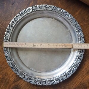 May include: A silver serving tray with an ornate, embossed design around the edge. The tray measures approximately 8 inches in diameter.
