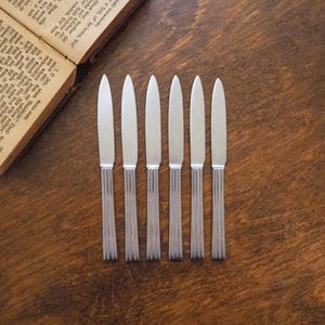 May include: Six silver dinner knives with a brushed finish and a slightly rounded tip. The knives are arranged in a row on a wooden surface.