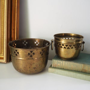 Könnte beinhalten: Zwei Messing-Blumentopf mit herzförmigen Ausschnitten. Der kleinere Topf hat zwei Griffe und steht auf zwei Büchern. Der größere Topf hat eine Aufhängeschlaufe.