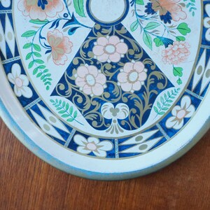 Puede incluir: Una bandeja de servir de metal blanco con un dise&ntilde;o floral azul y dorado. La bandeja tiene un borde azul claro y un borde festoneado.
