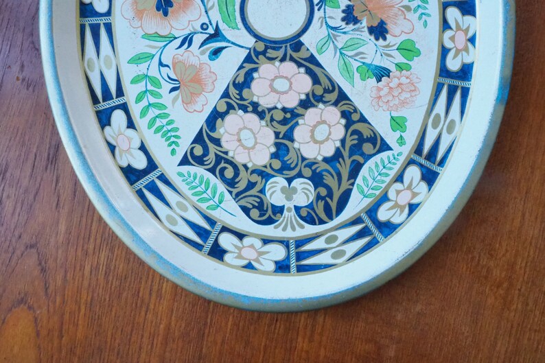 Puede incluir: Una bandeja de servir de metal blanco con un dise&ntilde;o floral azul y dorado. La bandeja tiene un borde azul claro y un borde festoneado.