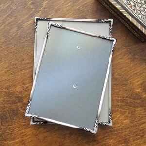 Puede incluir: Dos marcos de fotos rectangulares con un acabado plateado metálico y detalles decorativos de cuerda trenzada en los bordes. Los marcos están apilados ligeramente desplazados, cada uno con un tablero trasero gris claro liso con dos pequeños orificios circulares.