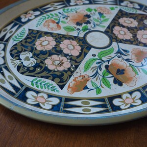 Puede incluir: Una bandeja de servir de metal vintage con fondo blanco y un dise&ntilde;o floral en tonos azules, verdes, rosas y naranjas. La bandeja tiene un borde dorado y un patr&oacute;n geom&eacute;trico azul y dorado alrededor del borde.
