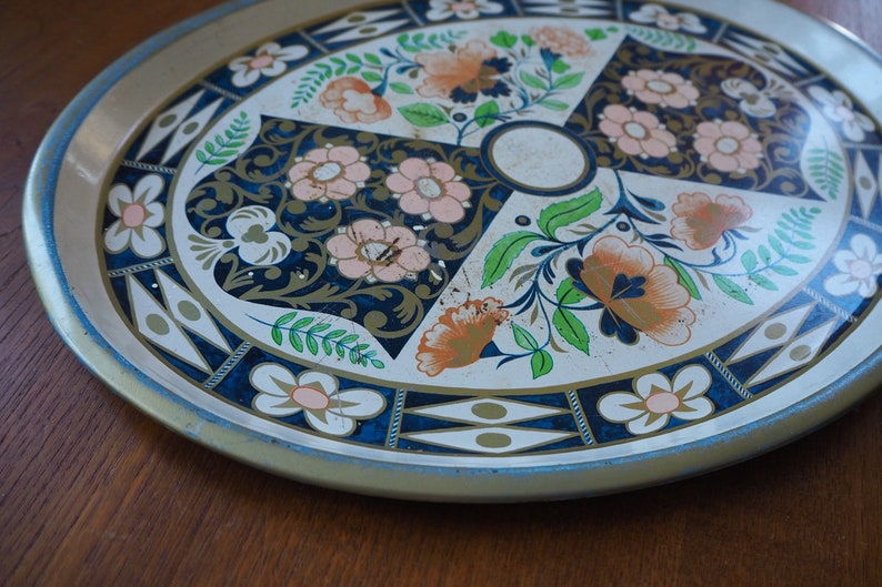 Puede incluir: Una bandeja de servir de metal vintage con fondo blanco y un dise&ntilde;o floral en tonos azules, verdes, rosas y naranjas. La bandeja tiene un borde dorado y un patr&oacute;n geom&eacute;trico azul y dorado alrededor del borde.