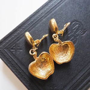 May include: A pair of gold-tone clip-on earrings featuring a detailed apple design. The earrings are made of metal and have a shiny finish.